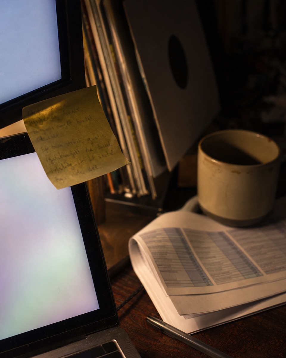 Maya's desk at night: a handwritten rule on a sticky note stuck to a monitor, beside vinyl records, a mug, and printouts.